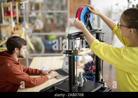 Junge weibliche Büroangestellte ändern Schieber mit roten Faden auf 3d Drucker mit Kollege Vernetzung im Hintergrund Stockfoto
