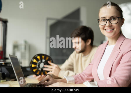 Hübsche junge Manager betrachten Sie mit toothy Lächeln beim Sitzen durch Arbeitsplatz vor Laptop mit Co-worker auf Hintergrund Stockfoto