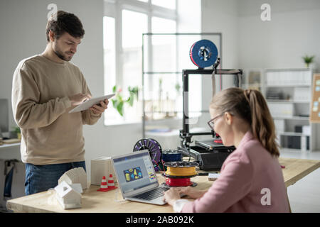 Junge Designer mit Touchpad online Arbeiten im Stehen vor 3d-Drucker und seine Kollegen die Skizze der Zahlen Stockfoto