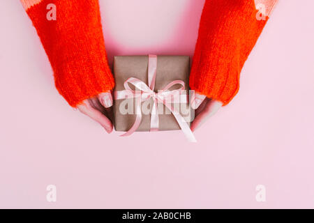 Die Frau Hände in warmen gestrickten Pullover mit Maniküre halten Geschenkbox mit Handwerk Papier und Pink Ribbon auf rosa Hintergrund gewickelt. Stockfoto