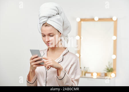 Junge sauber Frau in seidenpyjama Blättern durch Nachrichten in Smartphone Stockfoto