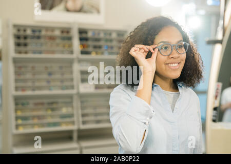 Hübsche Junge lächelnd brünette Frau, die versucht, auf neue Brillen vor Spiegel auf der Hintergrund der Anzeige mit Brillen Stockfoto
