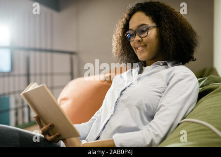 Glückliche junge kluge Studentin in neue Brille lesen Buch auf der Couch Stockfoto