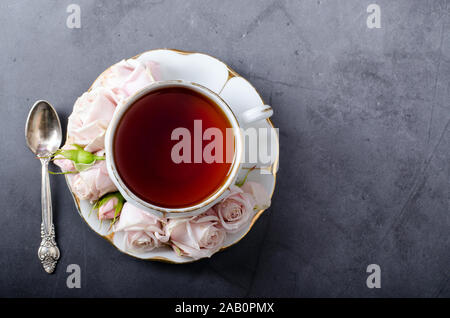 Tea time Kulisse. Top-down-Stillleben mit Vintage White Porzellan Tasse Tee mit sanftem Rosa Rosen und schön Kaffee Löffel auf einem dunkelgrauen Hintergrund. Roman Stockfoto