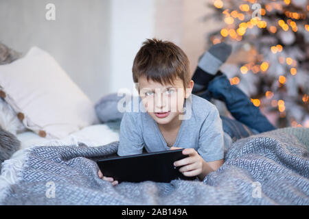 Ernsthafte junge Holding Tablet in Händen. Kind spielt Computerspiele auf Tablet. Junge liegt auf dem Bett gegenüber Weihnachtsbaum vor Weihnachten. Schwarzer Freitag und Stockfoto