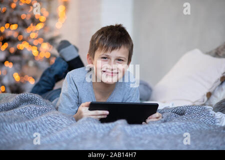 Gerne schöner Junge mit einem Tablett in den Händen. Kind spielt Computerspiele auf Tablet. Junge liegt auf dem Bett gegenüber Weihnachtsbaum vor Weihnachten. Schwarz Frida Stockfoto