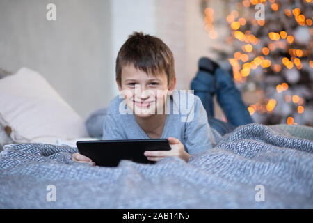 Gerne schöner Junge mit einem Tablett in den Händen. Kind spielt Computerspiele auf Tablet. Junge liegt auf dem Bett gegenüber Weihnachtsbaum vor Weihnachten. Schwarz Frida Stockfoto