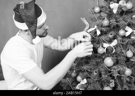 Mann in santa hut Weihnachtsbaum dekorieren mit Kugeln, Schleifen, Girlande. Weihnachten, Neujahr, Silvester, urlaub Vorbereitung, Feier. Festliche Dekorationen und Verzierungen Konzept Stockfoto