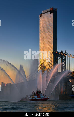 Ein Manhattan Square skyscraper mit der Manhattan Bridge und FDNY feuerlöschboot Wasserstrahlen bei Sonnenuntergang. Lower Manhattan, New York City, NY, USA Stockfoto