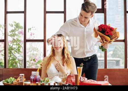 Lächelnd attraktiven jungen Mann mit Blumenstrauß aus frischen Rosen in der Hand, die Augen von seinem glücklichen Freundin am Tisch im Restaurant sitzen Stockfoto