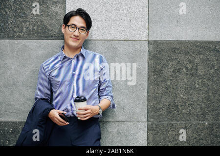 Portrait von Stattlichen lächelnden jungen asiatischen Mann mit Tasse Kaffee und Smartphone lehnte an der Wand und auf der Suche an Kamera Stockfoto