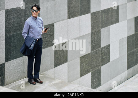Schöne elegante junge Mann, der draußen an der Wand und Texting beim Warten auf jemanden Stockfoto