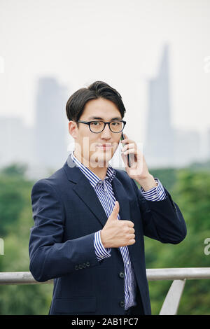 Lächelnd asiatische Geschäftsmann angezeigt Thumbs-up, wenn am Telefon sprechen mit Kollegen Stockfoto