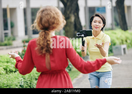 Junge asiatische Frau mit Smartphone angeschlossen und in die Hände der Handbewegung weibliche Blogger, dass sie weiter sprechen vor der Kamera zu zeigen, zu Einbeinstativ Stockfoto
