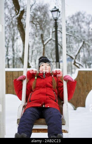 Winter Portrait eines kleinen Mädchens in einen Hut und eine rote Jacke auf einer Schaukel Stockfoto