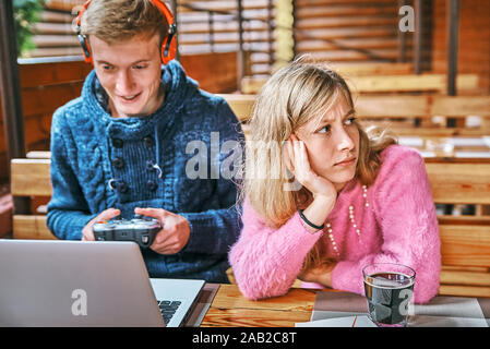 Jungen Kerl spielt Spiele bei einem Laptop in einem Cafe. das Mädchen in der Nähe ist ein Buch zu lesen. Ein Verschleiß Konzept Stockfoto