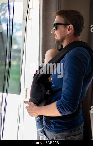 Papa schaut aus dem Eingang an einem sonnigen Tag, während sein Baby schläft ein Baby carrier. Stockfoto