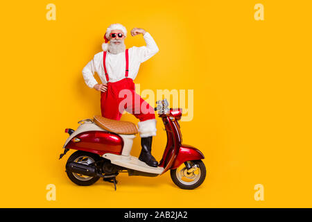 Voller Körper Foto von Santa ehrgeizige Mann stand in der Nähe von Moped zeigen Stärke Bizeps geliefert schweren Geschenke trendige Sun specs Hosen tragen cap Shirt isoliert Stockfoto