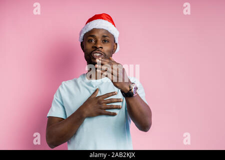 Gerne dankbar dunkelhäutigen jungen Mann im Gespräch mit Freund, tragen, Santa Hut, halten Sie eine Hand auf Herz, Gestik, über rosa Hintergrund posiert. Mittei Stockfoto