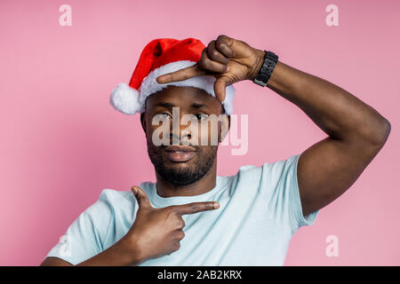 Nahaufnahme der Ruhe schwere unrasiert afro-amerikanische Mann, Rahmen mit beiden Händen, legere weisses T-Shirt, Santa Hut, stehend gegen Rosa b Stockfoto