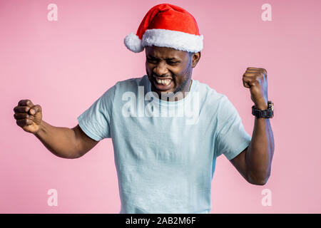 Wütend, wütend afrikanischer Mann ballte die Fäuste und Zähne, Hände, tragen t-shirt, Santa Hut, Pläne für Weihnachten nicht wahr, auf Rosa posing b Stockfoto