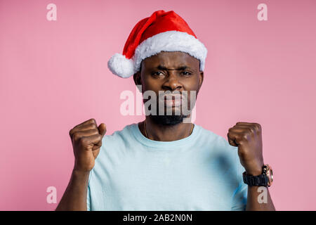 Wütend, wütend afrikanischer Mann die Hand, ballte die Fäuste und Kamera mit Verärgerung, t-shirt, Santa Hut, auf rosa Hintergrund posiert. Stockfoto