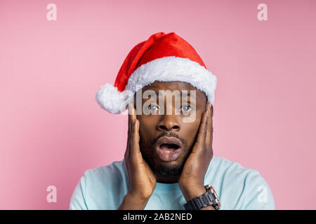Closeup Schuss attraktive dunkelhäutige Mann mit Weihnachtsmütze schaut mit surprisement, sieht etwas Betäuben, hält die Handflächen auf Wangen, ständigen Gege Stockfoto
