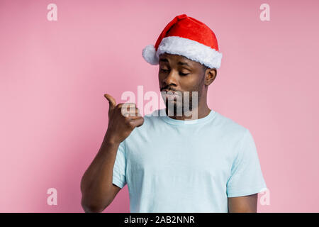 Horizontale Schuß von schweren Bärtigen dunkelhäutigen Mann, Santa Hut, weißes T-Shirt und zeigte mit dem Daumen zur Seite, gegen Pink Studio an der Wand stehen. F Stockfoto