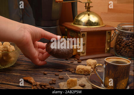 Die Hand des Mädchens hält einen Marshmallow in Schokolade, einer Tasse Kaffee, Kaffeebohnen, brauner Zucker, Kaffeemühle auf einer hölzernen Hintergrund Stockfoto