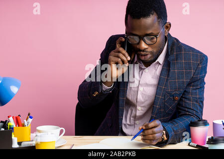 Ernsthafte afro-amerikanische Geschäftsmann in formale Abnutzung, Gläser, Gespräch am Handy, bei notepad suchen, der die Feder, sitzen am Schreibtisch im Büro. Job, b Stockfoto