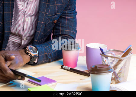 Close up 7/8 Schuß des dunkelhäutigen Geschäftsmann, tragen, Klage, stilvolle Armbanduhr, sitzen am Schreibtisch aus Holz, Smartphone. Büroartikel, Schale Stockfoto