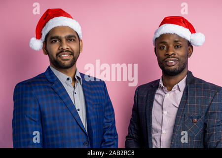 Nahaufnahme von zwei Multiethnischen attraktive zuversichtlich Unternehmer tragen Anzüge, Nikolausmützen, lächelnd über rosa Hintergrund isoliert. Weihnachten Stockfoto