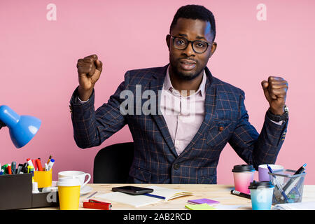 Junge afrikanische amerikanische Geschäftsmann, Hände, ballte die Fäuste,, am Arbeitsplatz im Büro zu sitzen, Erfolge feiern, über Rosa backgroun isoliert Stockfoto