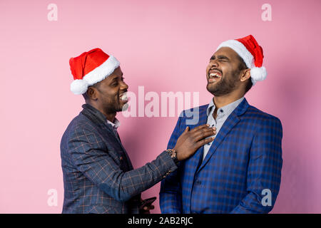 Freudige afro-amerikanische Unternehmer und ein indischer Geschäftsmann zu lächeln und zu lachen, über rosa Hintergrund stehen, tragen Nikolausmützen. Beste Freunde in Stockfoto