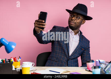 Stilvolle schöne afro-amerikanische Unternehmer am Schreibtisch im Büro sitzen, Brille, Anzug, schwarzer Hut, Smartphone, lächelnd, wobei selfie. F Stockfoto