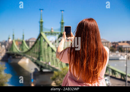 Junge rothaarige Mädchen in ein rosa Kleid macht ein Foto von der Brücke auf ein Smartphone an einem sonnigen Sommertag Stockfoto