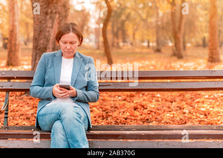 Frau mit Mobiltelefon auf Pausen während der Arbeitszeit beim Sitzen auf einer Parkbank an sonnigen Herbsttag Stockfoto