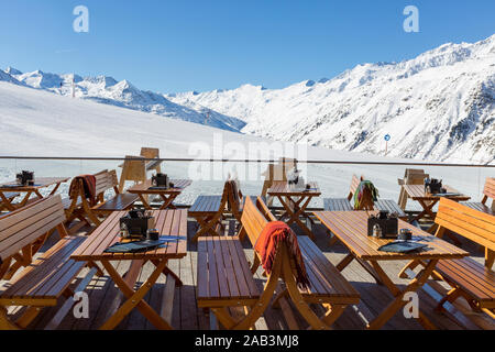 Stühle und Tische auf eine leere Terrasse an einem Ski cafe in einem österreichischen Resort mit schneebedeckten Bergen hinter Stockfoto