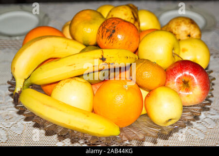 Fruchtmischung auf einer Glasplatte auf einem Tisch mit weißer Tischdecke aus Spitze, einschließlich Äpfel, Bananen, Orangen, Mandarin. Stockfoto