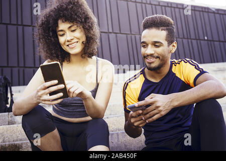 Glücklich athletische Latein Frau und ein junger Mann in Schwarz auf der Suche der Sport app in ihre Mobiltelefone auf der Treppe während der Rest im Training, Technik in Stockfoto