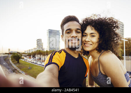Selfie mit einem lächelnden Latin Frau und stattlichen schwarzen Mann während der Rest im Training durch die Stadt in der Dämmerung, Handy, Technik im Sport ein Stockfoto