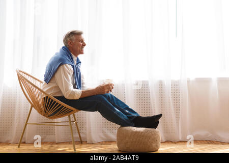 Älterer Mann auf Stuhl in der Nähe der Fenster und trinken Kaffee Stockfoto