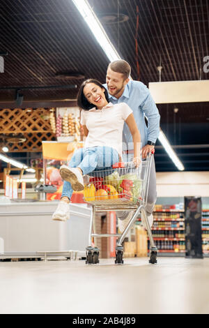 Die täglichen Einkäufe. Paar im Supermarkt zusammen Mann mit Karre, während Frau sitzt im Inneren Spaß fröhliche Lachen Stockfoto