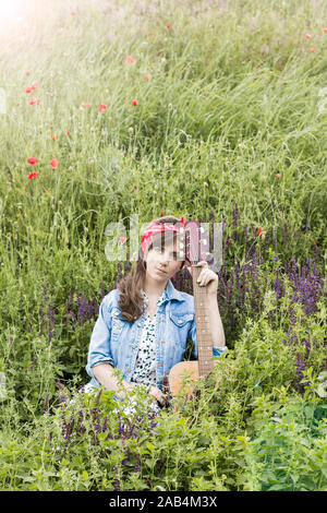 Schöne Mädchen mit einer Gitarre in Mohn. Junge Frau auf dem Gras. Ein Jugendlicher in einer Jeansjacke und eine rote Binde. Retro Stil Stockfoto