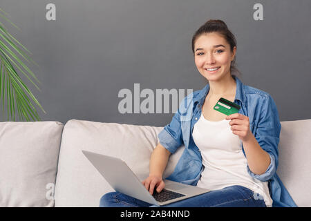 Online Shopping. Junge Frau sitzt auf einem Sofa studio isoliert auf Grau mit Kreditkarte mit Laptop lächelt Fröhlich Stockfoto