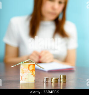 Frau mittleren Alters mit Modell Haus aus Israel Währungen neue israelische Schekel auf hölzernen Tisch. Frau Entspannung zu Hause, Gebäude Haus aus Israel 100 und 50 Banknoten. selektive Fokus auf Geld. Stockfoto