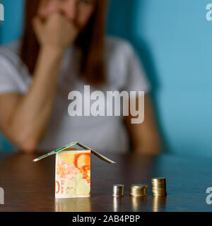 Frau mittleren Alters mit Modell Haus aus Israel Währungen neue israelische Schekel auf hölzernen Tisch. Frau Entspannung zu Hause, Gebäude Haus aus Israel 100 und 50 Banknoten. selektive Fokus auf Geld. Stockfoto