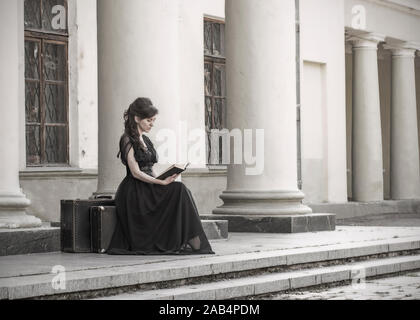 Schöne Mädchen in schwarz Abendkleid sitzen, ein Buch zu lesen. Ein Mädchen sitzt mit Koffern in der Nähe ein altes Gebäude mit Säulen. Schöne Frau mit Narben Stockfoto