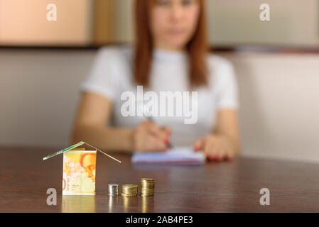 Frau mittleren Alters mit Modell Haus aus Israel Währungen neue israelische Schekel auf hölzernen Tisch. Frau Entspannung zu Hause, Gebäude Haus aus Israel 100 und 50 Banknoten. selektive Fokus auf Geld. Stockfoto