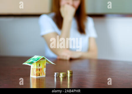 Frau mittleren Alters mit Modell Haus aus Israel Währungen neue israelische Schekel auf hölzernen Tisch. Frau Entspannung zu Hause, Gebäude Haus aus Israel 100 und 50 Banknoten. selektive Fokus auf Geld. Stockfoto